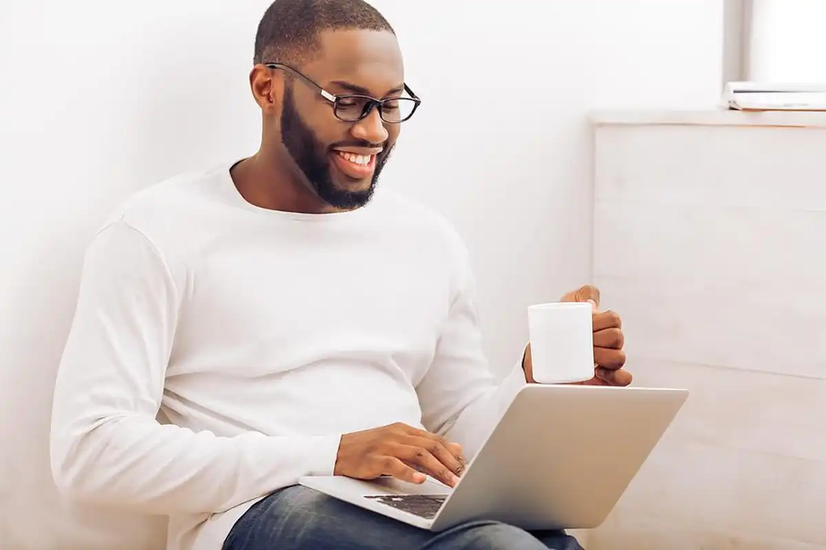 Man in a white shirt, wearing glasses, smiling while using a laptop and holding a white mug, seated in a bright room.